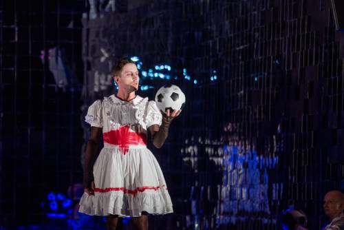 Aktor ubrany w białą sukienkę z czerwonymi dodatkami trzyma w ręku piłkę do piłki nożnej.